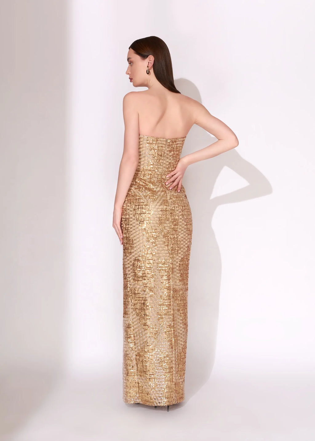 A woman modeling a strapless, floor-length gold sequined gown from the back, standing against a plain white studio background.