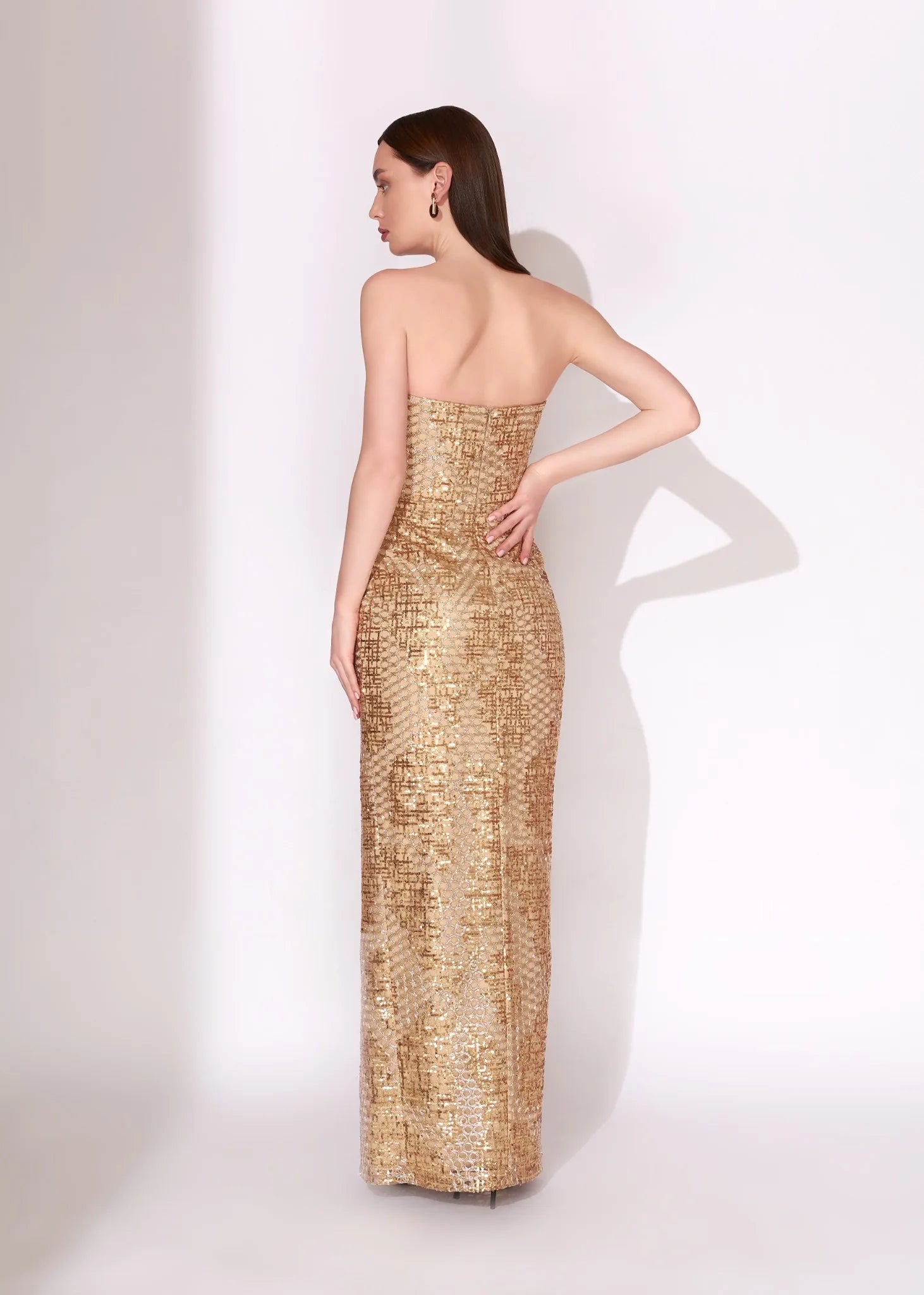 A woman modeling a strapless, floor-length gold sequined gown from the back, standing against a plain white studio background.