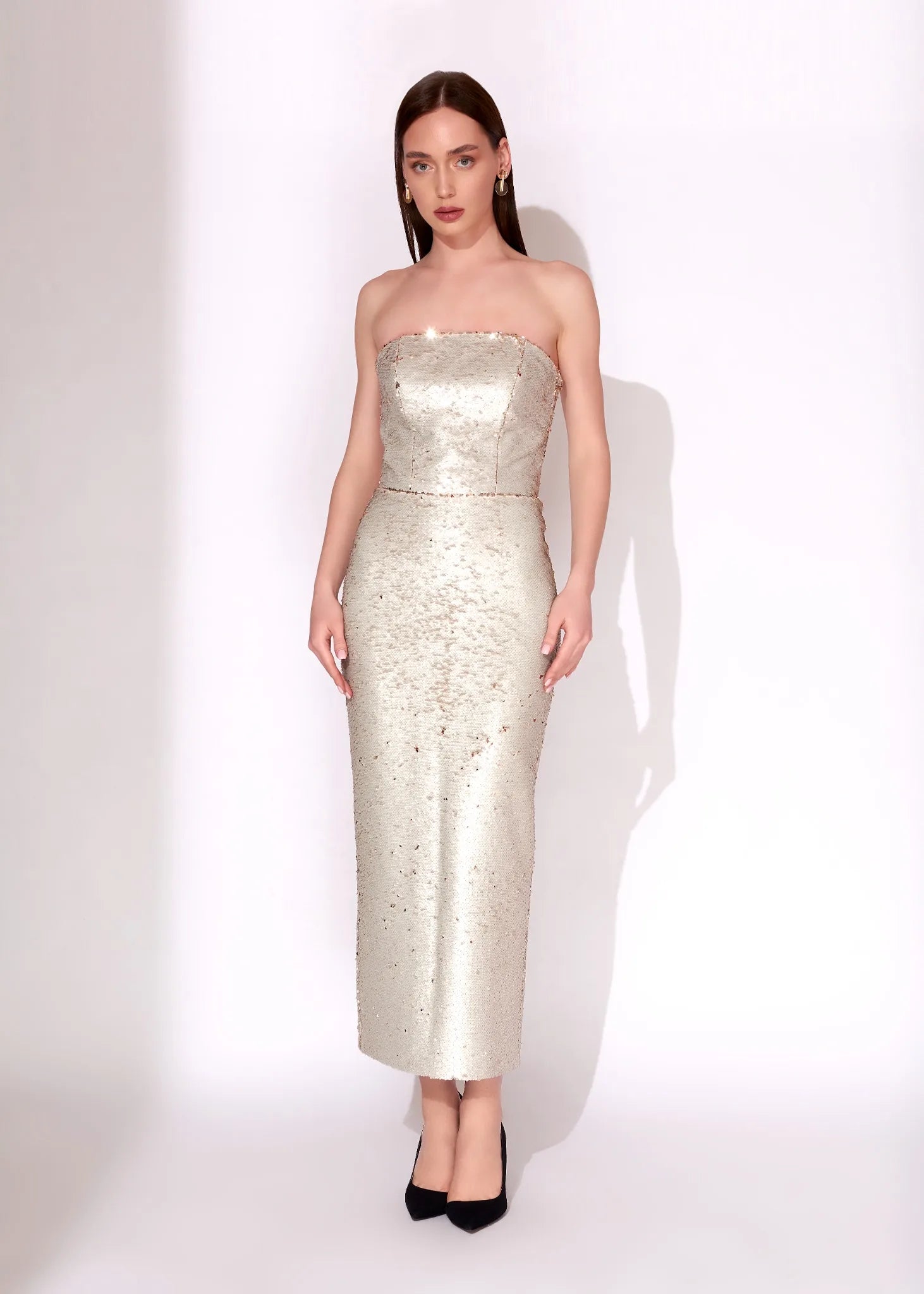 A woman modeling a strapless, floor-length champagne-colored sequined gown with black pointed-toe heels, standing against a plain white studio background.